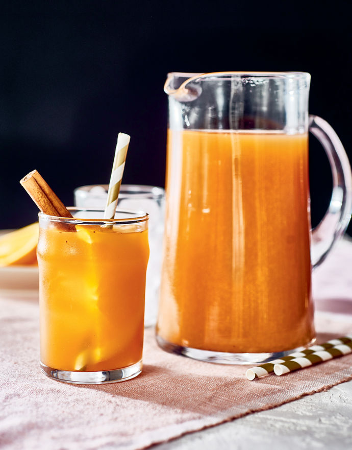 Jug of orange iced tea with a glass, holding a stirrer and cinnamon stick