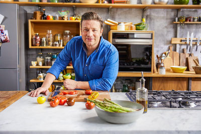 Jamie wearing a blue shirt, leaning on the kitchen counter with chopped tomatoes and asparagus in front of him