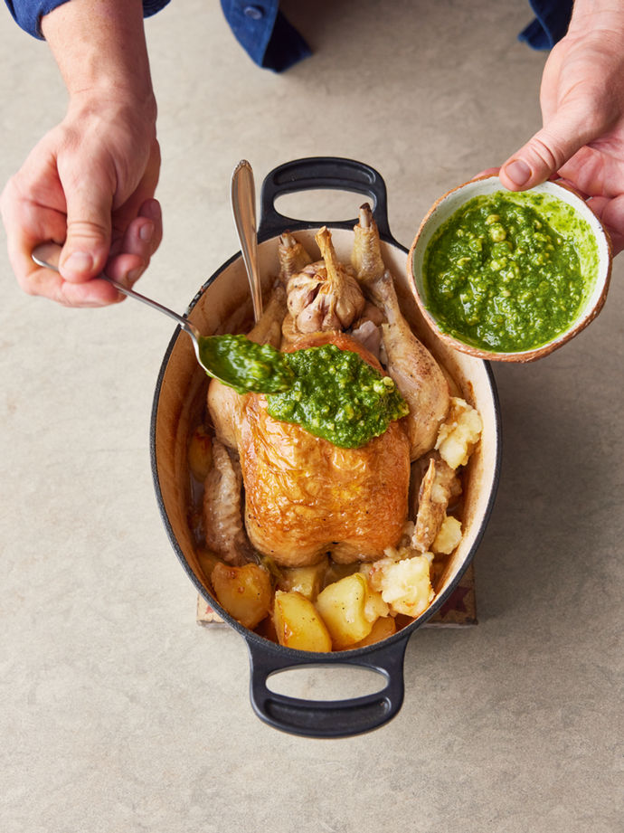 Whole roast chicken in a pot with potatoes and pesto drizzled on top